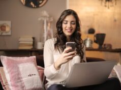 woman sitting on sofa while looking at phone with laptop on lap